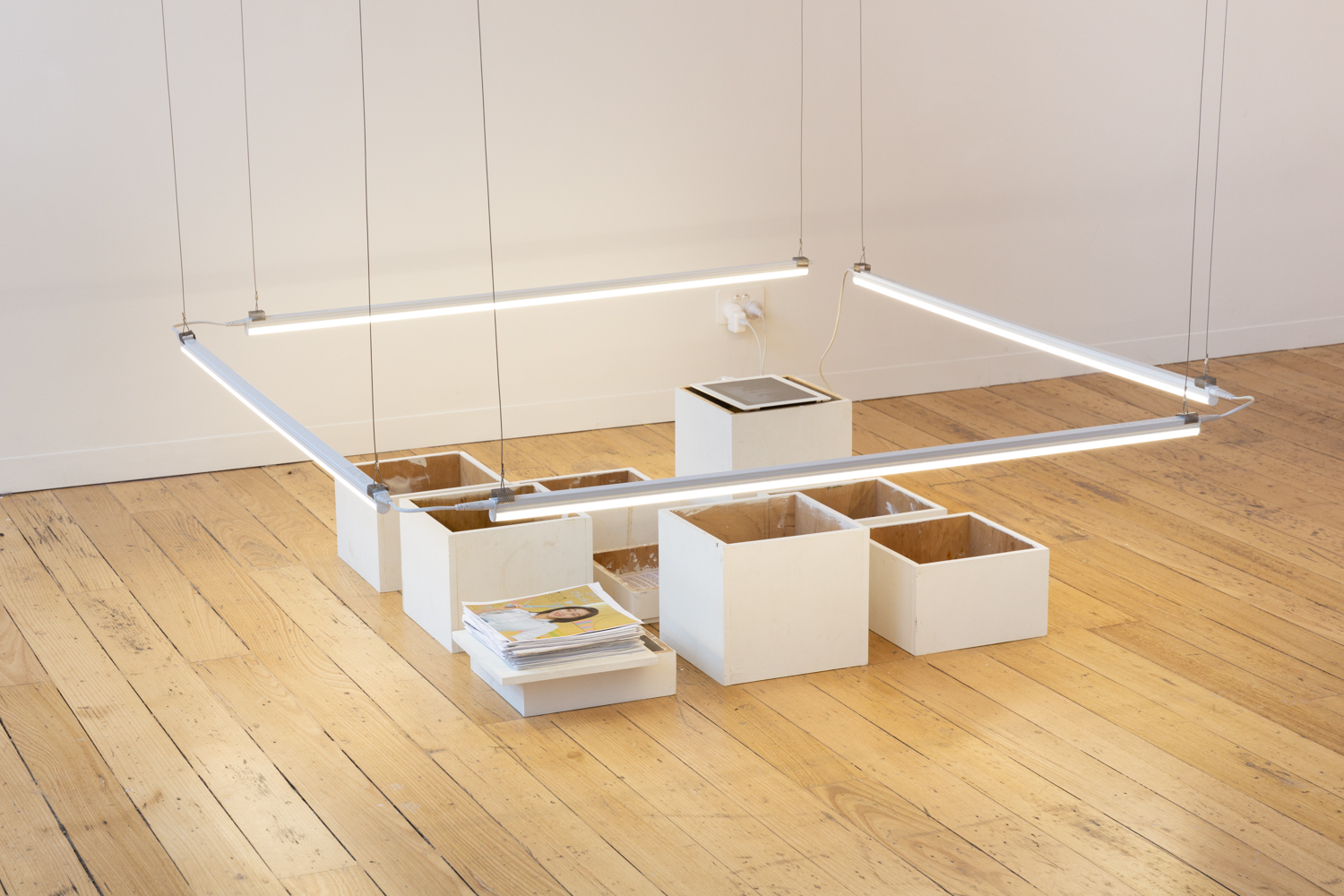 A series of 9 plinths of varying heights placed together to create a rectangle. On top of each plinth is an artwork. The plinths are surrounded by low hanging lights.
