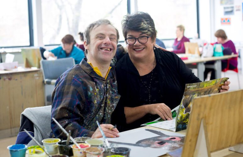 Sue Roff and artist Michael Trasancos are seated at a desk inside the Arts Project Australia studio.