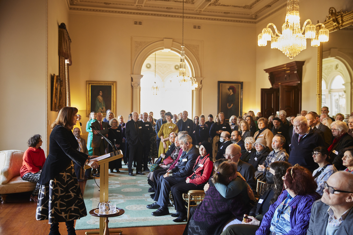 A large crowd of people are seated inside Government House. They are facing the Governor who is delivering a speech from behind a lectern.