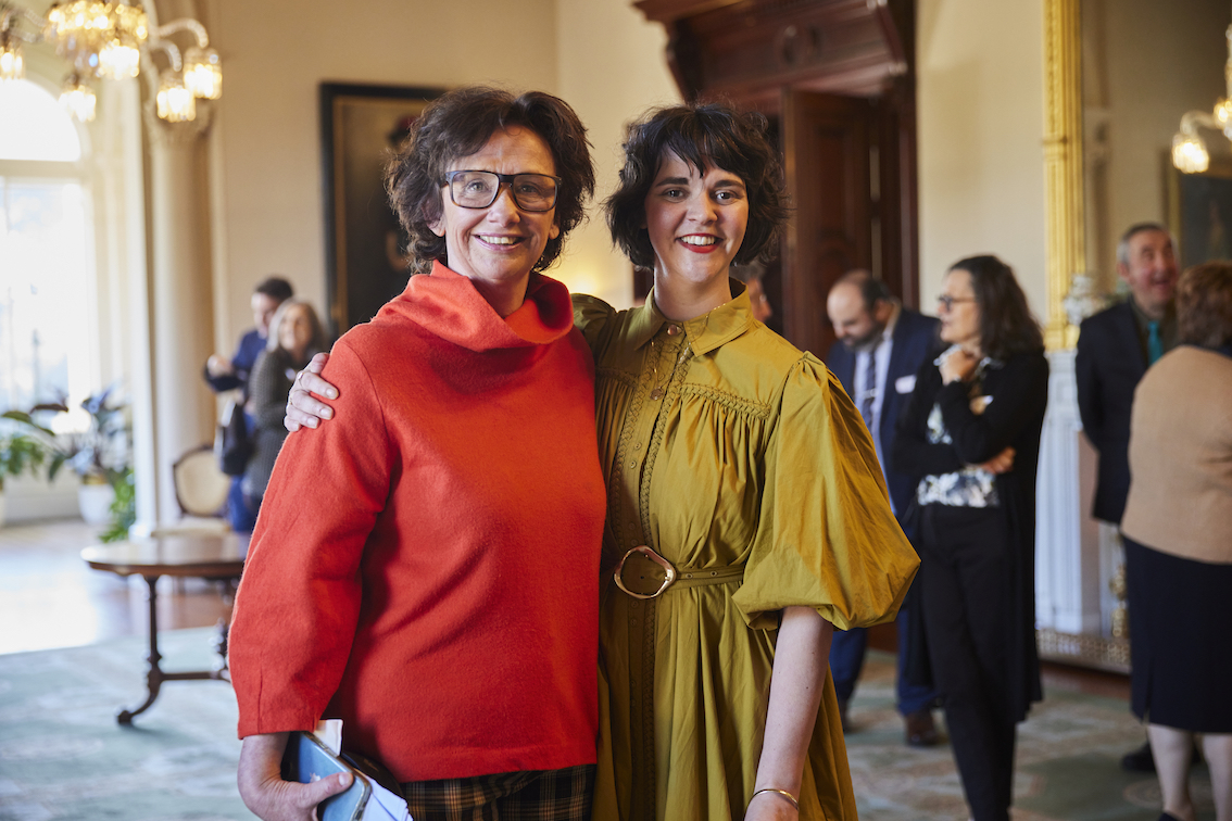 Two people are smiling and facing the camera. They have their arms around each others shoulders.