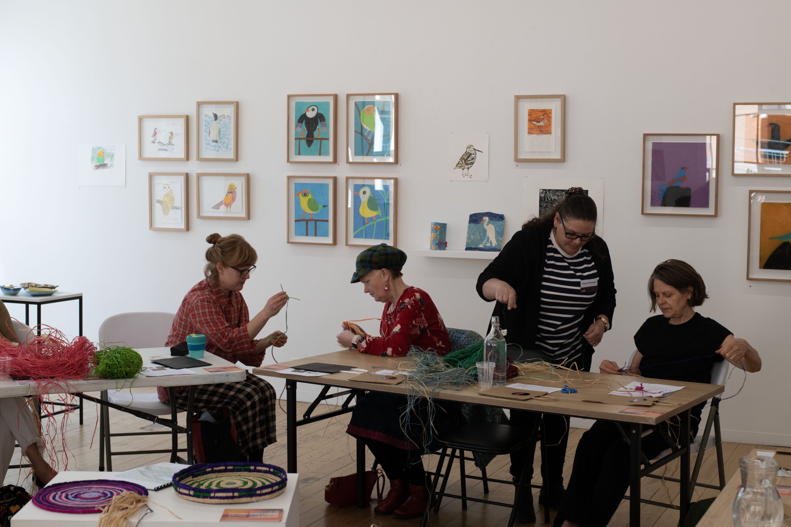 Three people are gathered around a table in the APA gallery. The person in the left corner is showing the two participants how to weave a basket. They are seated in front of a wall displaying a number of multi-coloured paintings and ceramics of birds.