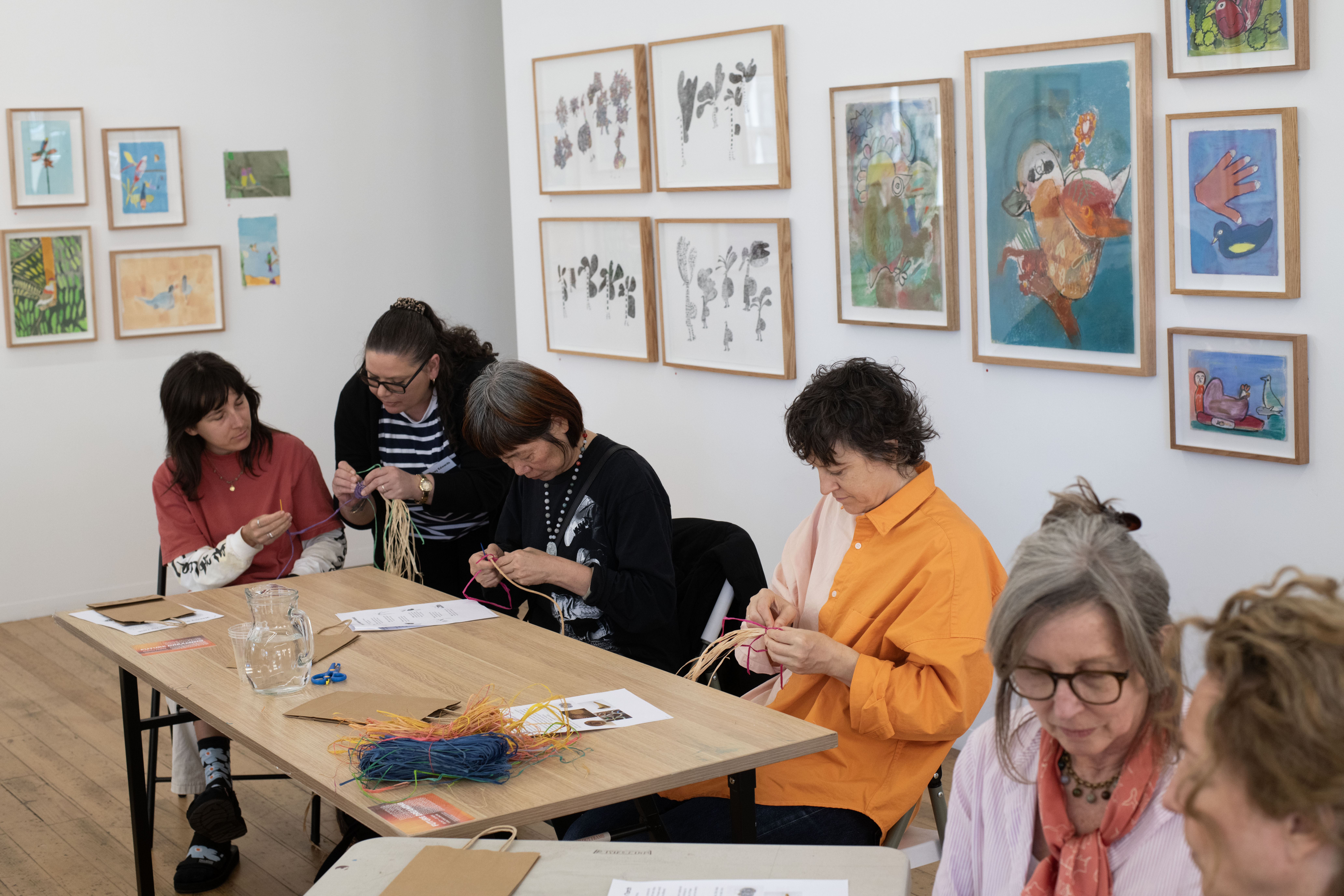 Participants are seated at their tables in the APA gallery. They are all engaged with their basket weaving projects.