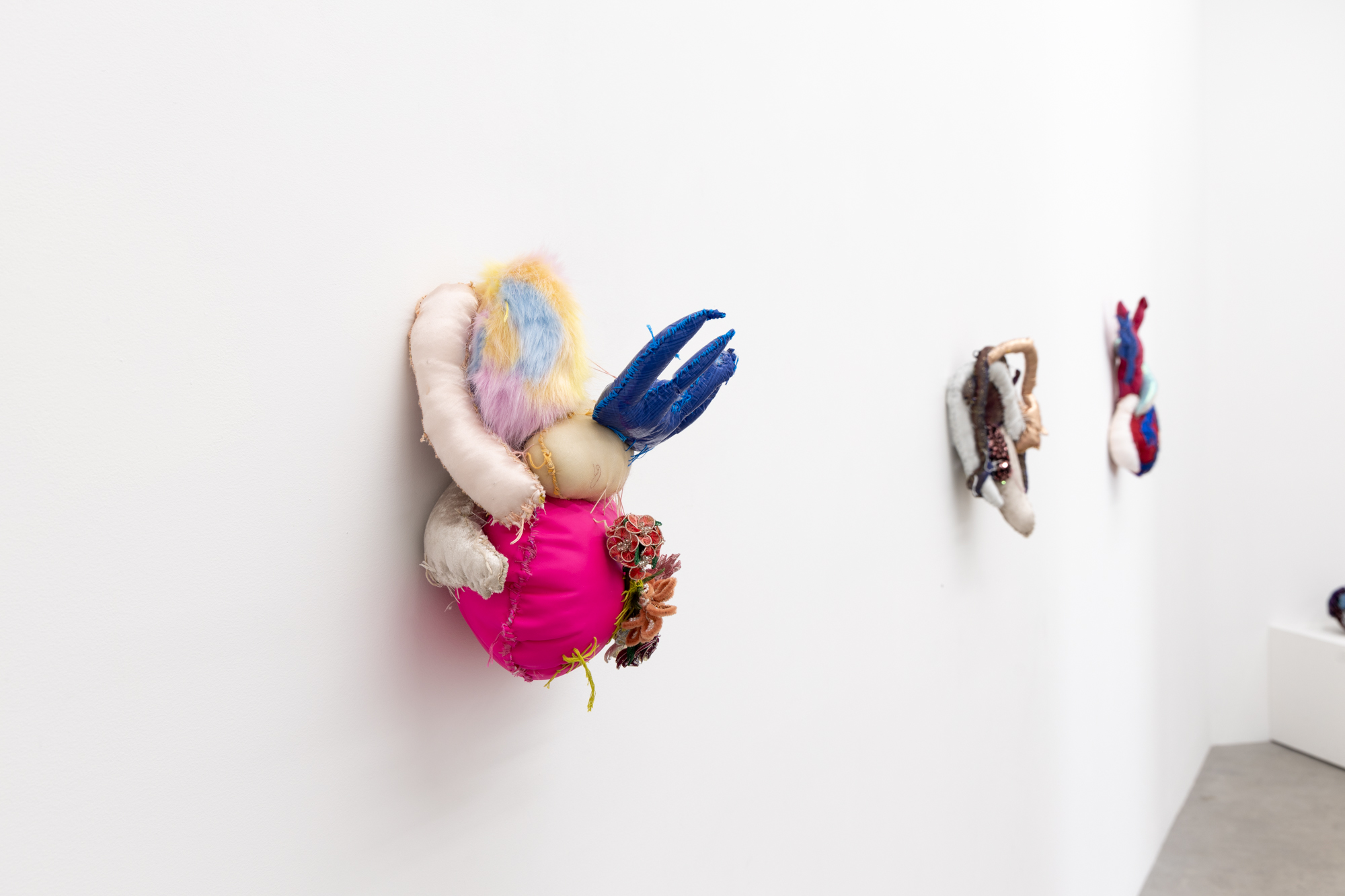 A photo of a soft sculpture made by Bronwyn Hack. The soft sculpture depicts a heart and is made of brightly coloured and textured fabrics with beads and thread woven between the different materials.