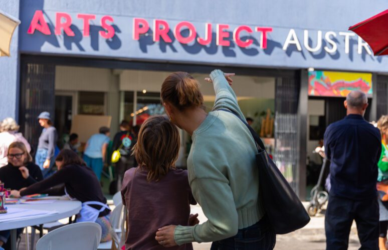 This is an image of two people standing out the front of the entrance to the APA studio. They have their back to the camera. The person on the right has one hand placed on the other person's back and is pointing towards the Arts Project Australia sign.
