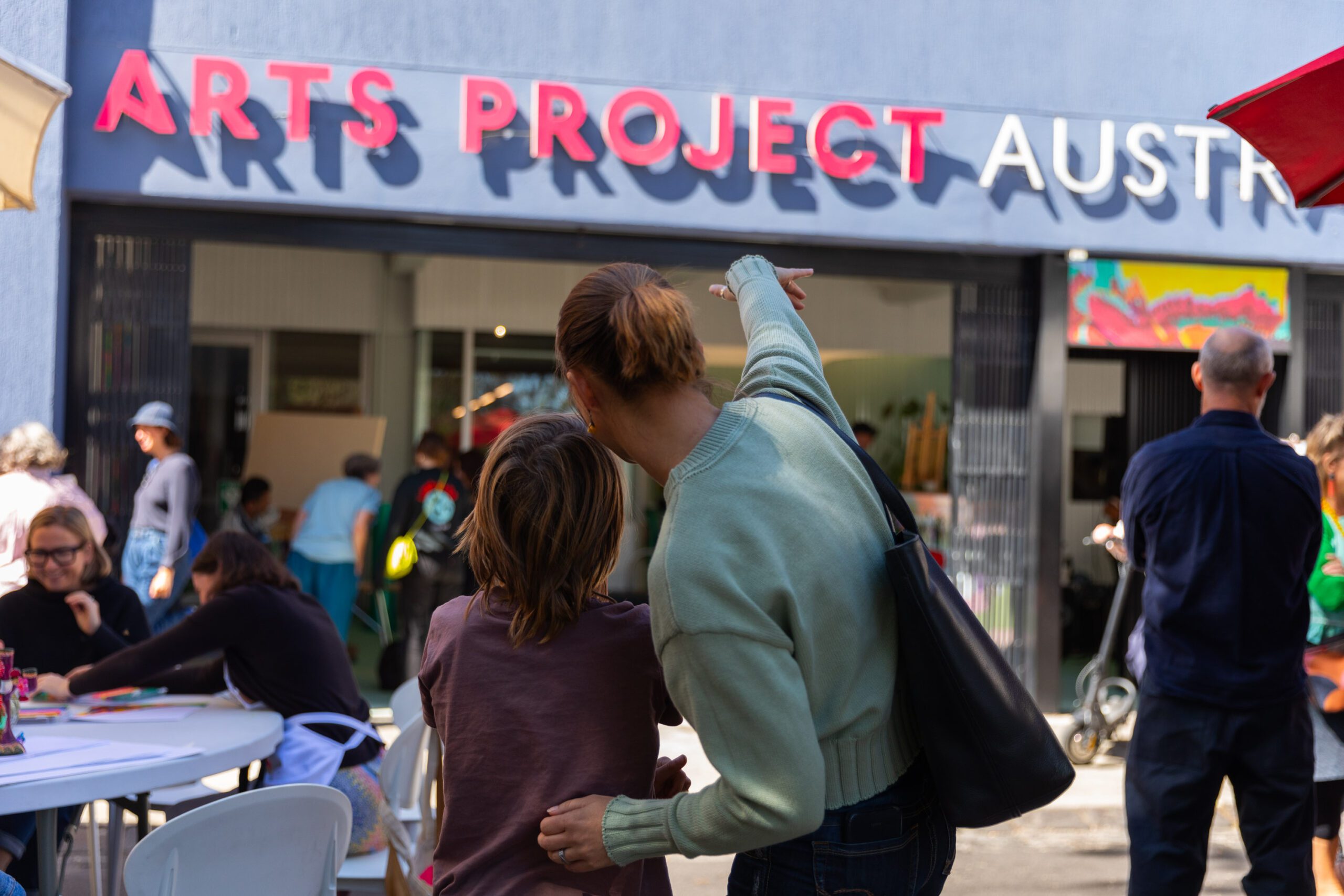 This is an image of two people standing out the front of the entrance to the APA studio. They have their back to the camera. The person on the right has one hand placed on the other person's back and is pointing towards the Arts Project Australia sign.