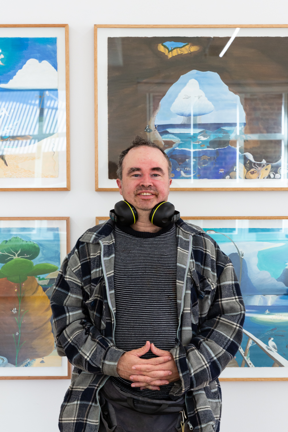This is a photo of Miles Howard-Wilks standing in front of his artworks in the APA gallery. He is smiling at the camera.