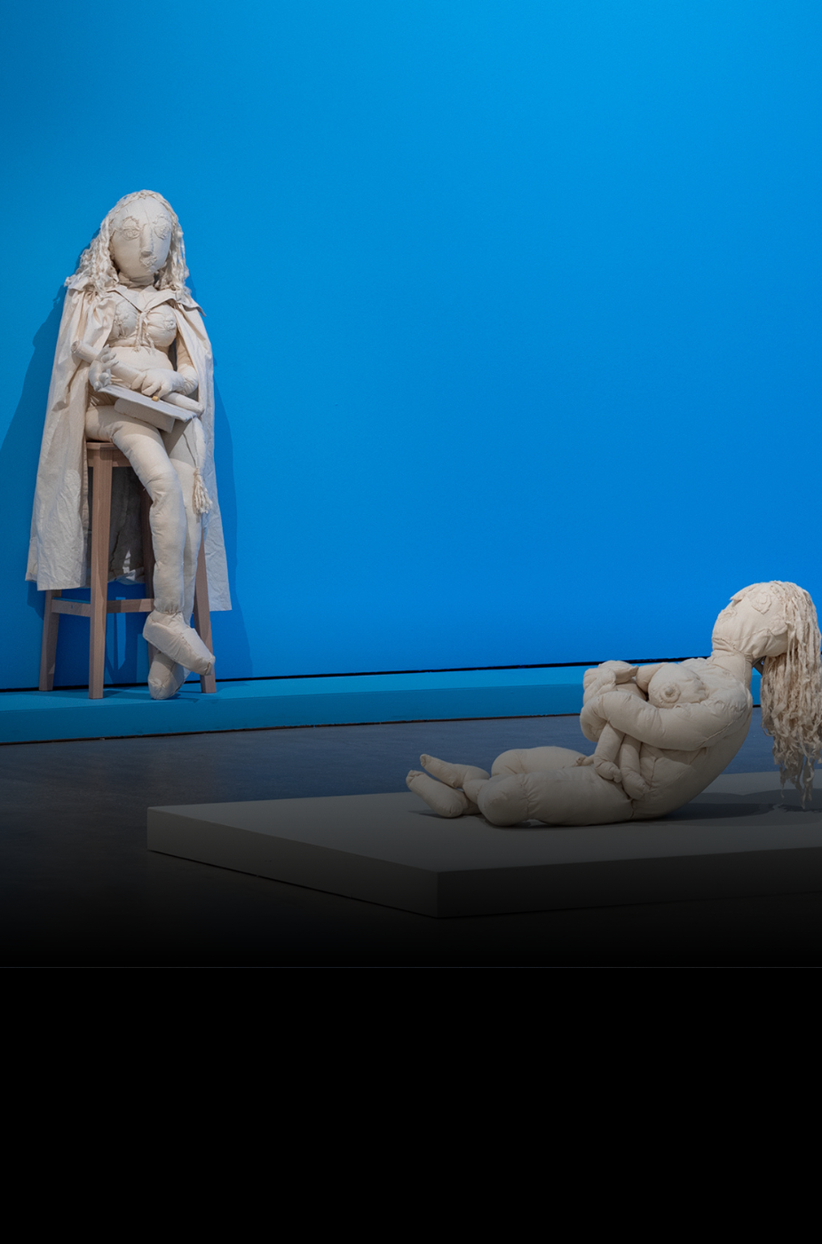 This is a photo of Mark Smith's soft sculpture works on display in a museum. The works depict three people. On the left a person is reading a book. In the centre a person is nursing a child and on the right a person is on a swing. They are placed against a blue wall.