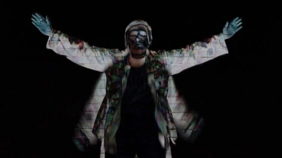 This is a photo of Max Cheevers. He is wearing a white coat covered in paint. His arms are up in the air.