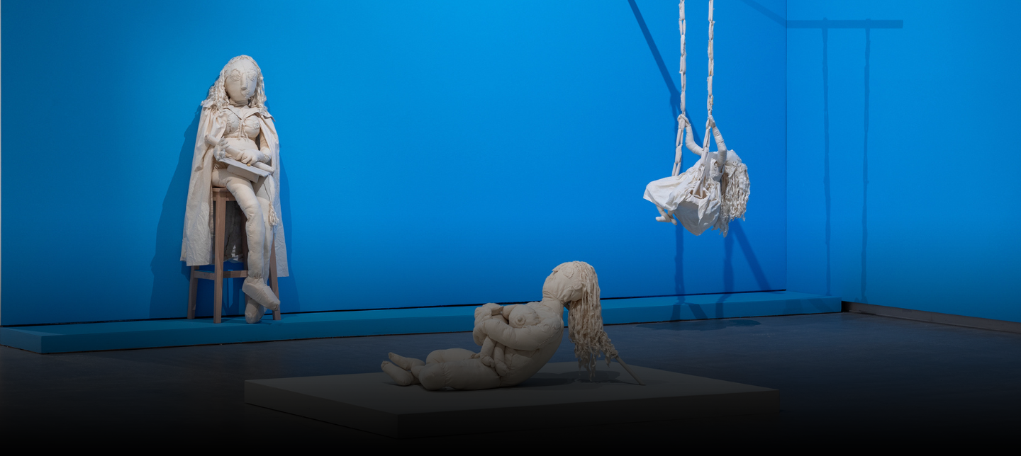 This is a photo of Mark Smith's soft sculpture works on display in a museum. The works depict three people. On the left a person is reading a book. In the centre a person is nursing a child and on the right a person is on a swing. They are placed against a blue wall.