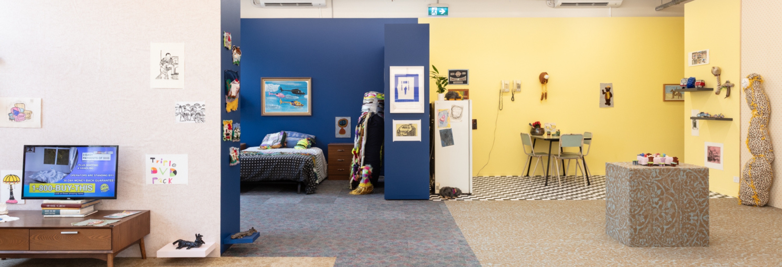 This image is a photo of the exhibition install for Houses, Spouses, Crises and Chrises. It shows an open plan house inside the APA gallery. There are blue walls in the bedroom. Yellow walls in the kitchen. Each room has its own patterned flooring. Artworks are displayed on and around the furniture.
