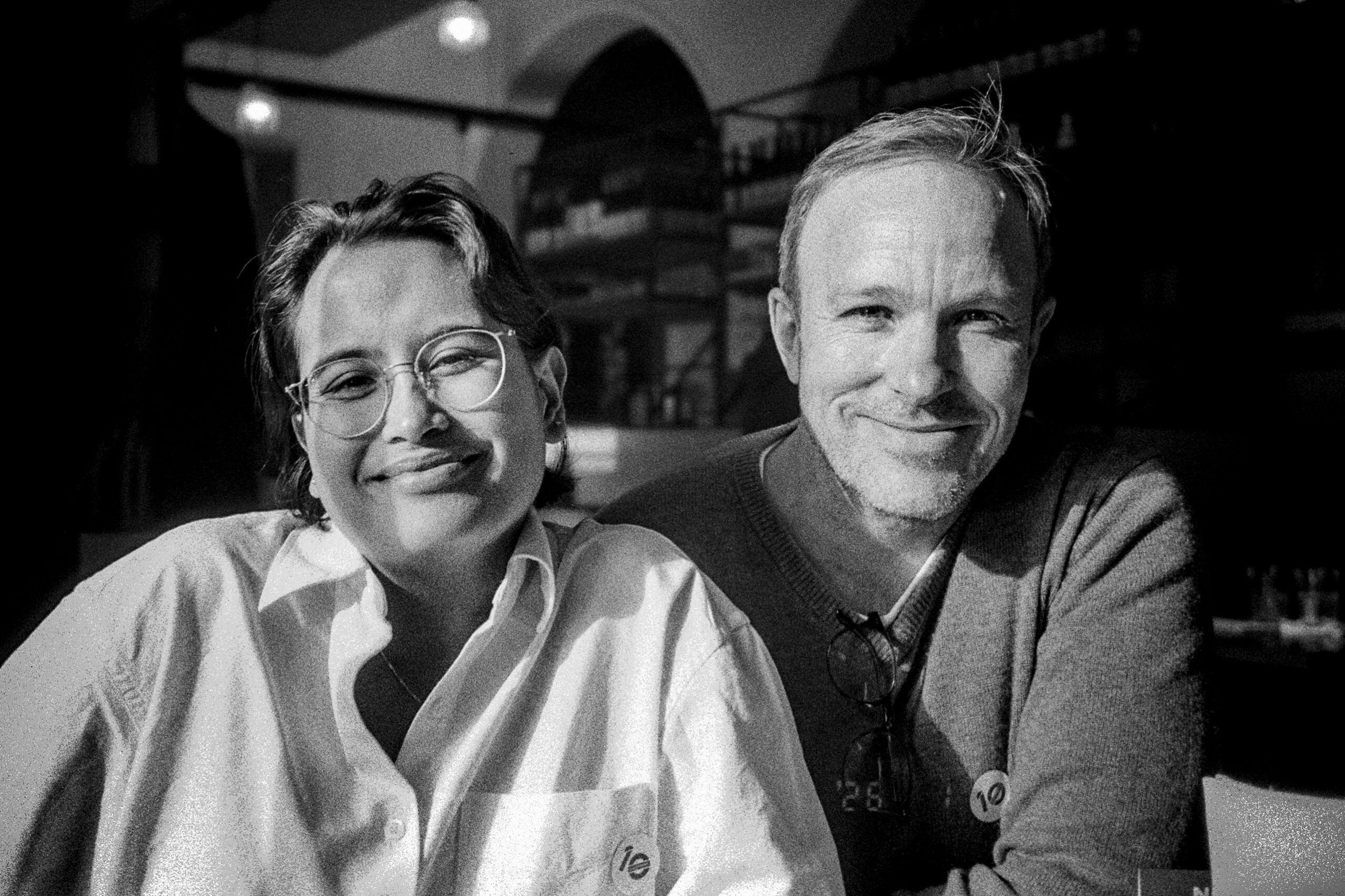 This is a black and white photo of two people smiling at the camera.